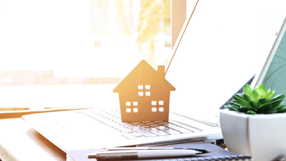 Miniature wooden house standing on a laptop, white flower pot, pens in bright sunlight
