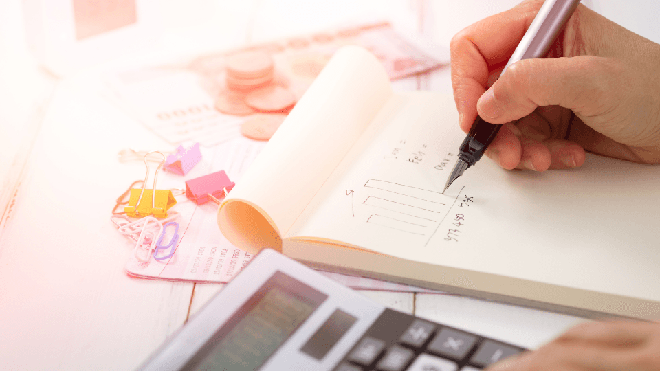 Somebody drawing a graph in notebook with calculator, staples. coins and bills