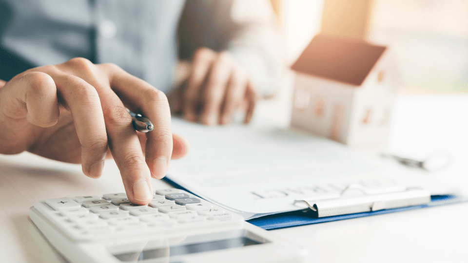 Man holds a pen and counts on a calculator With paper and house model on a desk