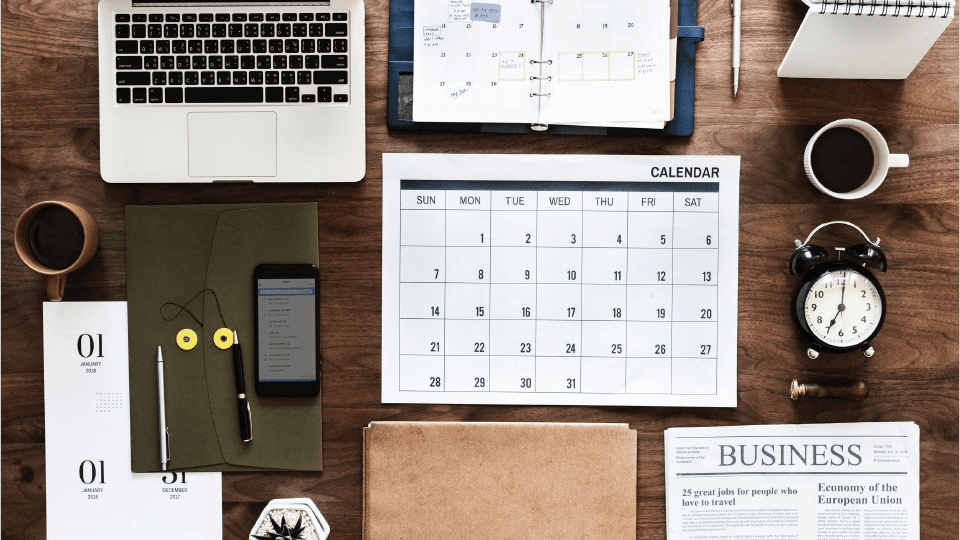 Lot's of things organized on the desk: laptop, notebooks, calendar, iphone, pens, clock, mugs, etc