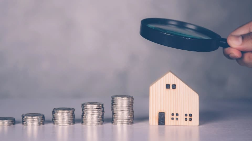 stack of coins and person holding a magnifying glass on a wooden house
